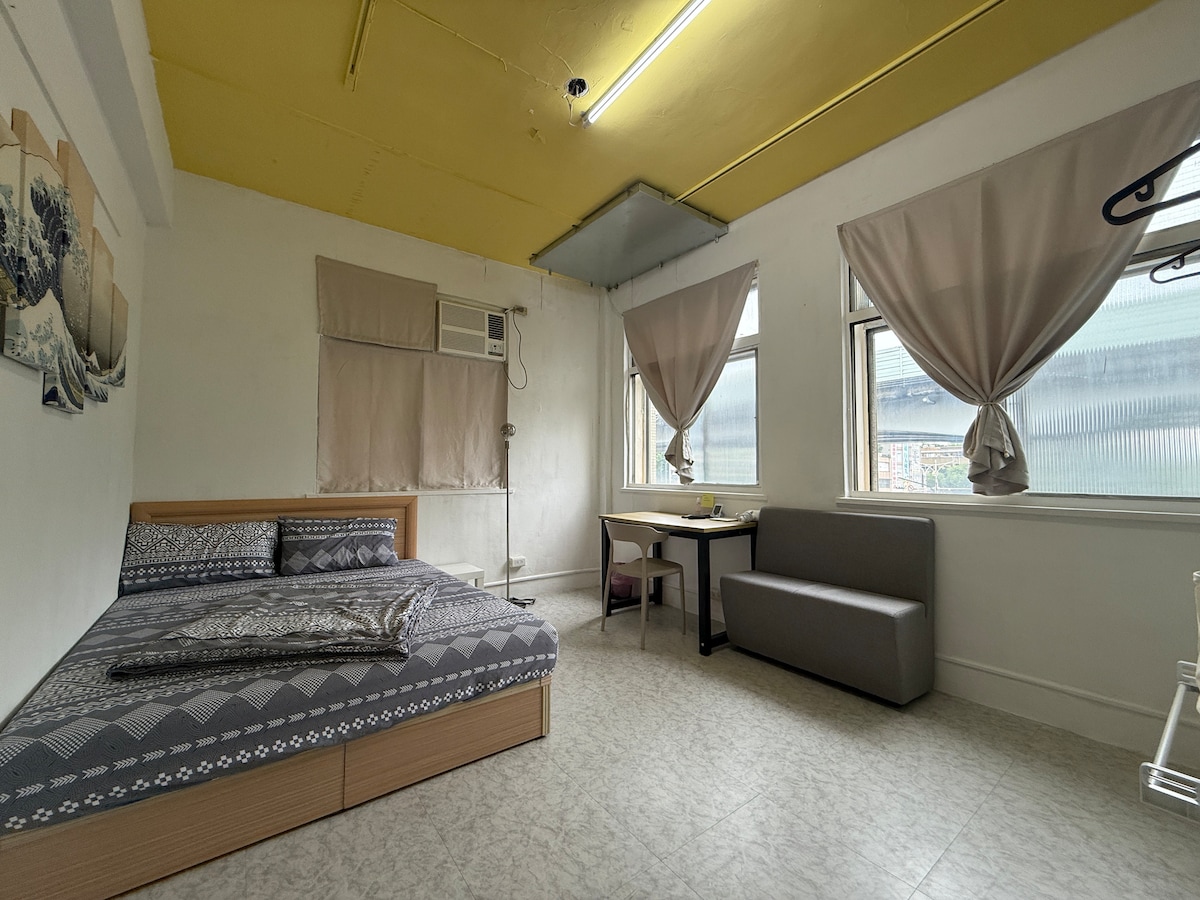 A minimalist room features a spacious bed with a patterned duvet and throw pillows. Light-filtering curtains frame large windows, allowing natural light to brighten the space. A small dining table and chairs complement a gray sofa, creating an inviting atmosphere for relaxation.