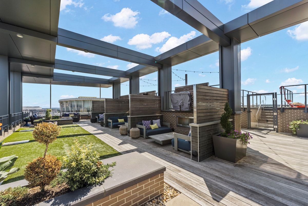 An expansive rooftop terrace features a mix of grassy areas and wooden decking. Comfortable seating arrangements are situated under a canopy of overhead beams. Planters with greenery add a touch of nature, enhancing the open and airy feel of the space.