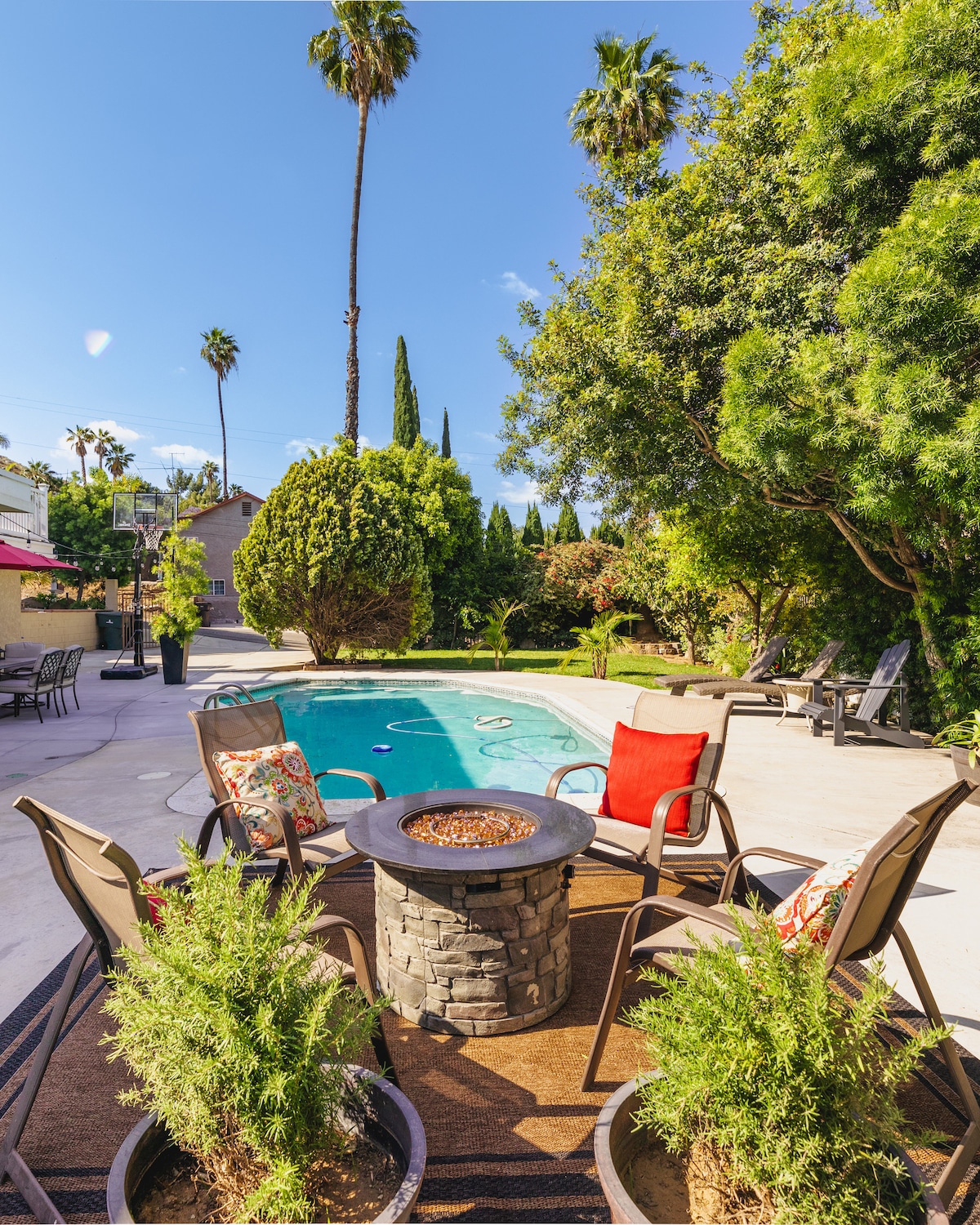An outdoor space showcases a pool surrounded by lush greenery and palm trees. A fire pit is surrounded by four chairs, providing a cozy gathering area. Various lounge chairs are positioned nearby, inviting relaxation in the sun.