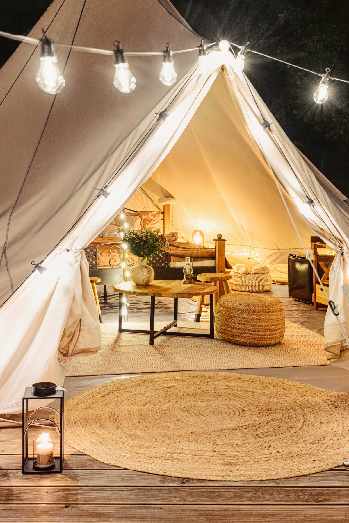 A fully equipped glamping tent is showcased, featuring warm lighting from string lights. Inside, a cozy seating area is accompanied by a round area rug and a lantern, while a small table and comfortable seating enhance the inviting atmosphere.