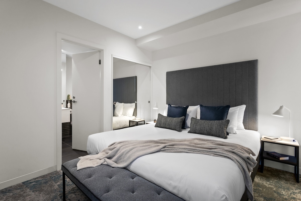 The sleeping area features a spacious bed with a gray upholstered headboard and layered bedding. A bench at the foot of the bed adds functionality. The room is well-lit, showcasing a minimalist design with a large mirror reflecting the contemporary style.