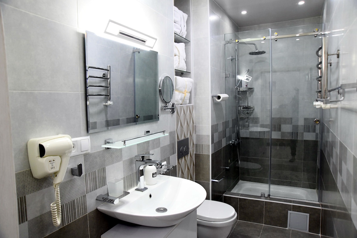 A modern bathroom featuring a glass shower enclosure and a stylish sink with a mirror. Towels are neatly arranged on a shelf above. Fixtures are in a silver tone, complementing the gray tiles that create a sleek appearance. A hairdryer is mounted on the wall for convenience.