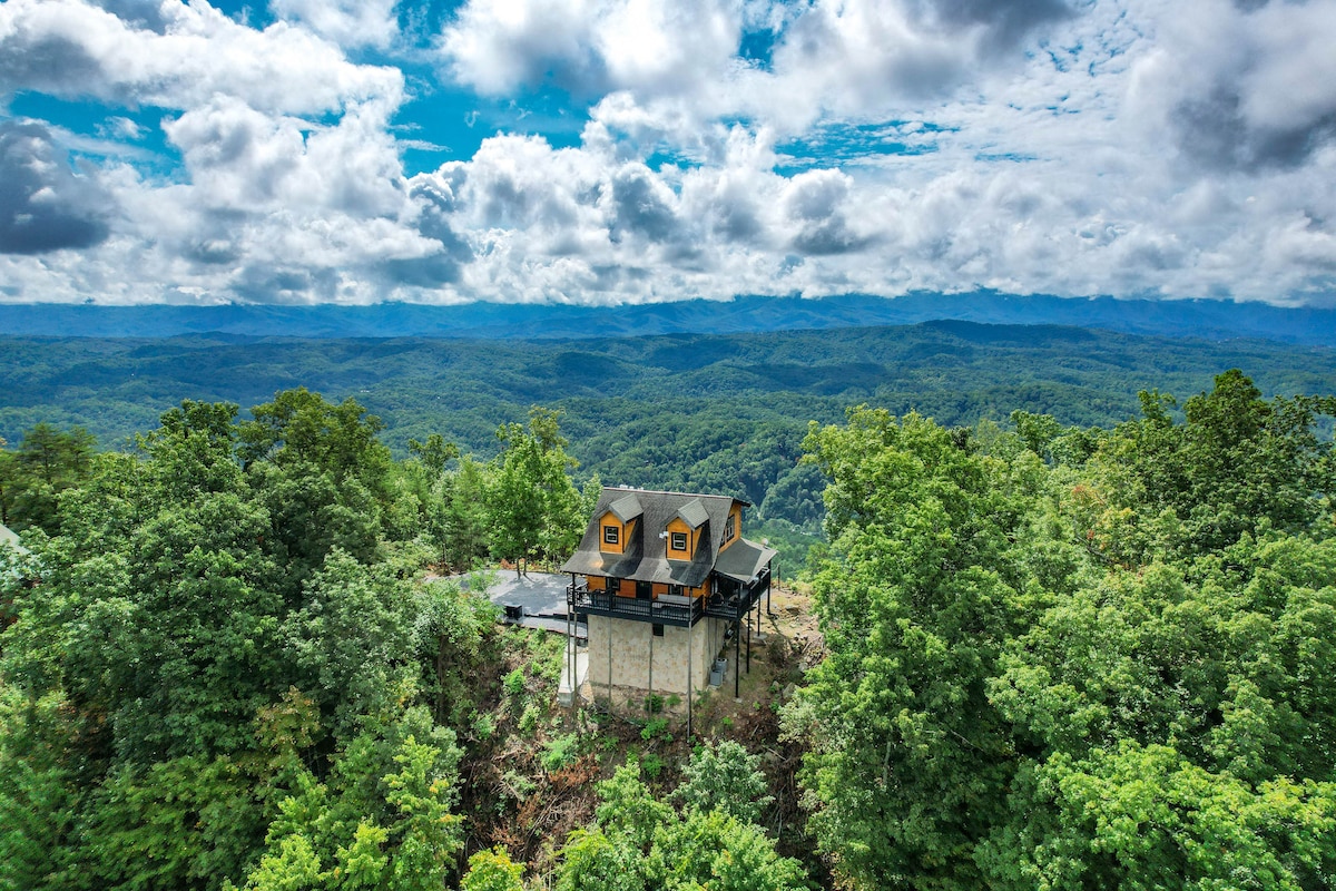 A mountaintop cabin is set among lush greenery, showcasing a dual-sided view of rolling hills and forests. The structure features large windows and a unique elevated design, surrounded by the natural beauty of the landscape and a wide expanse of sky above.