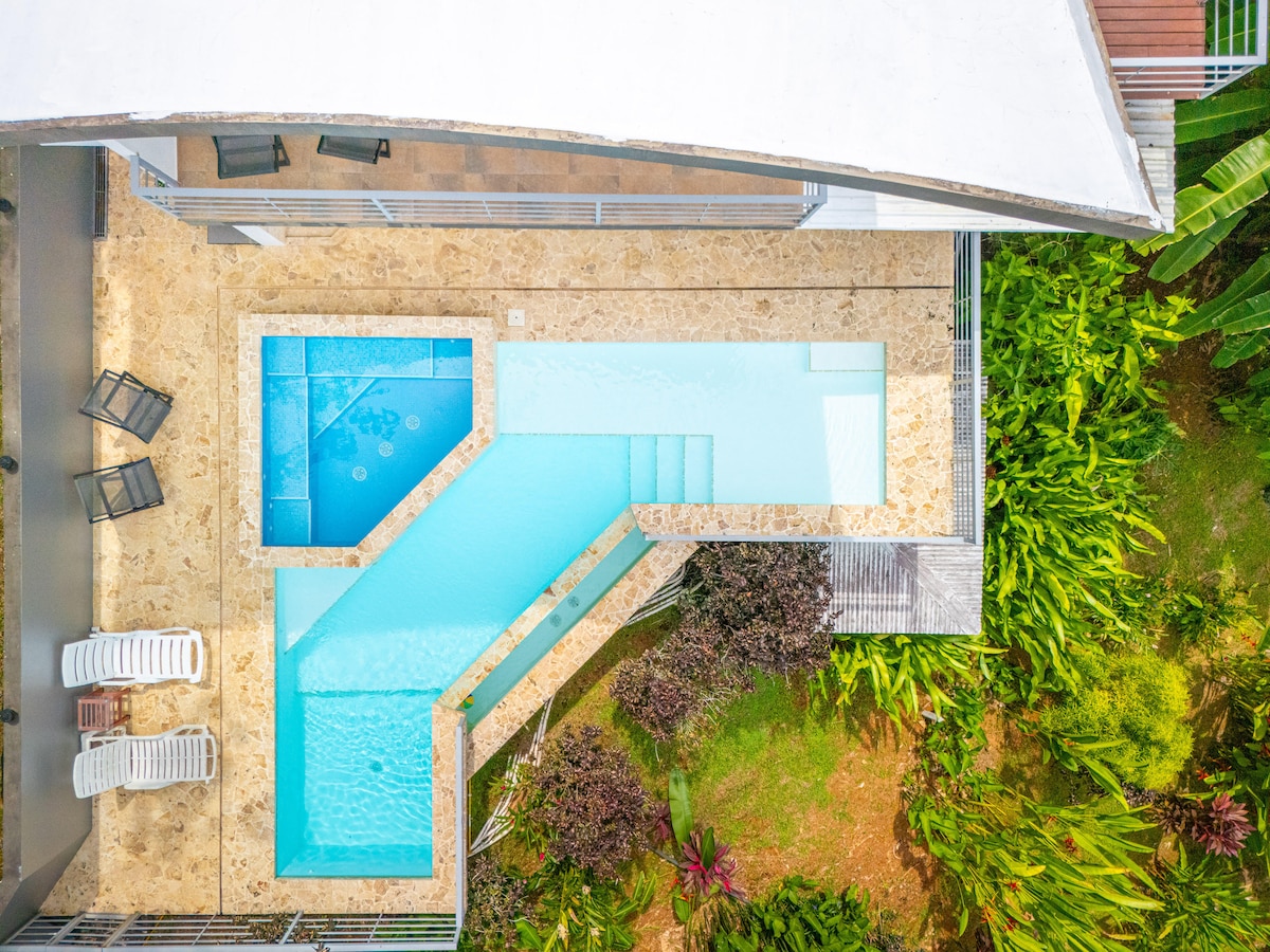 An aerial view of a modern infinity-edge pool is showcased, with clear blue water seamlessly blending into the surrounding outdoor space. The pool features varying depths and is bordered by a beige stone deck. Lush greenery and tropical plants surround the area, enhancing the serene environment.