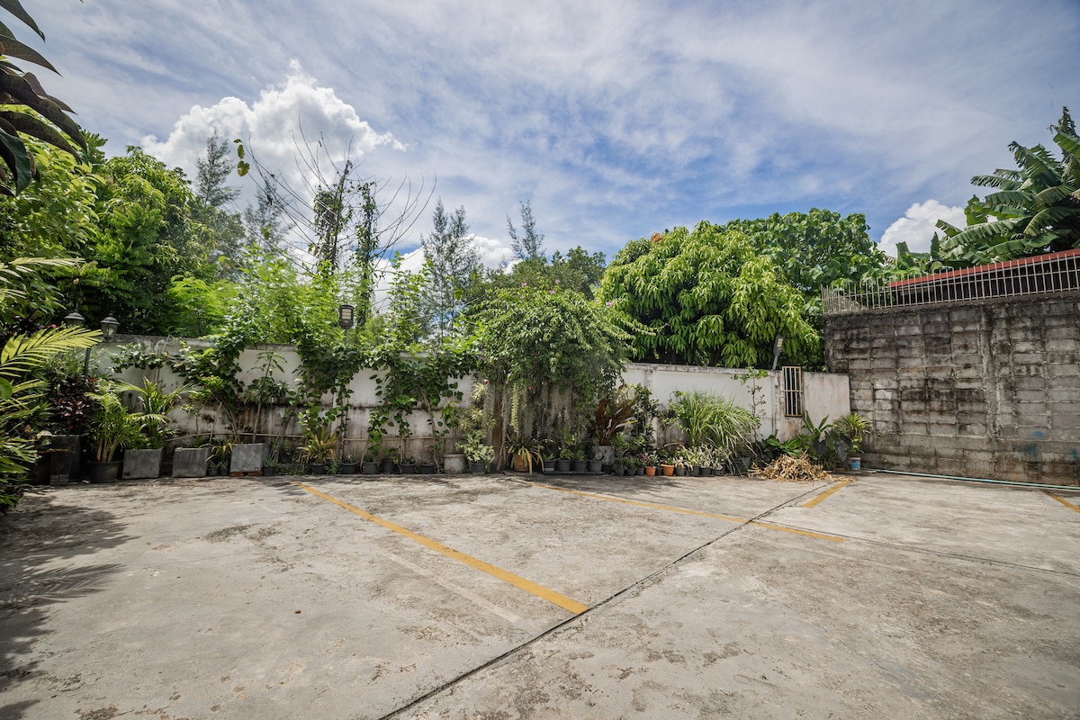An outdoor parking area is surrounded by lush greenery, featuring various potted plants along the perimeter. The concrete ground is marked with yellow lines, providing clear parking spaces. The sky is partly cloudy, adding a natural ambiance to the scene.