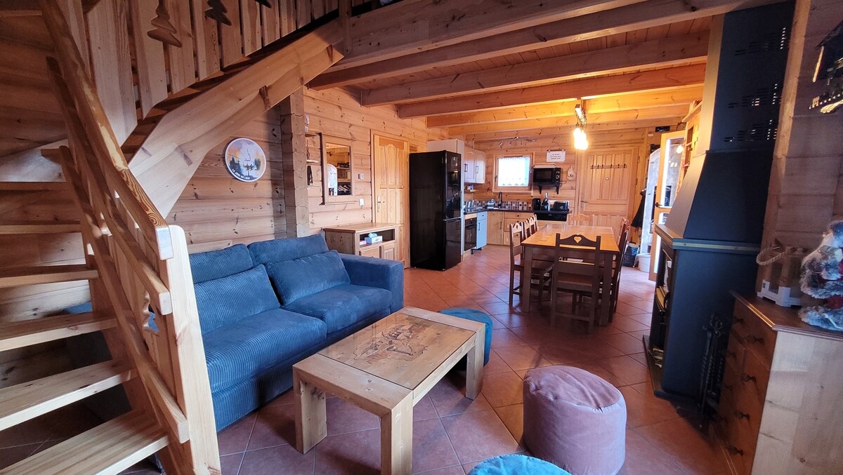 A spacious living area, featuring wooden walls and warm lighting. A comfortable sofa sits beside a wooden coffee table, with dining table and chairs nearby. The kitchen is equipped with modern appliances, including a black refrigerator and oven. A staircase leads to the upper level.