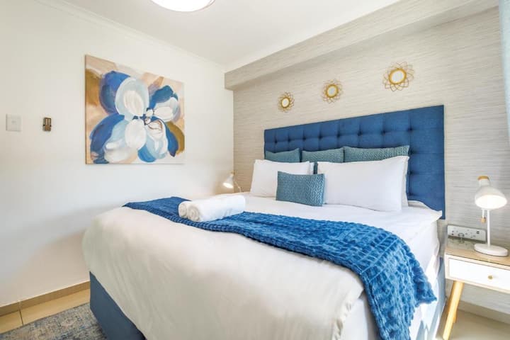 Unwind in this serene bedroom—featuring a tufted blue headboard, crisp white linens with a navy throw, ambient bedside lighting, and golden wall décor that adds a touch of luxury to your stay.