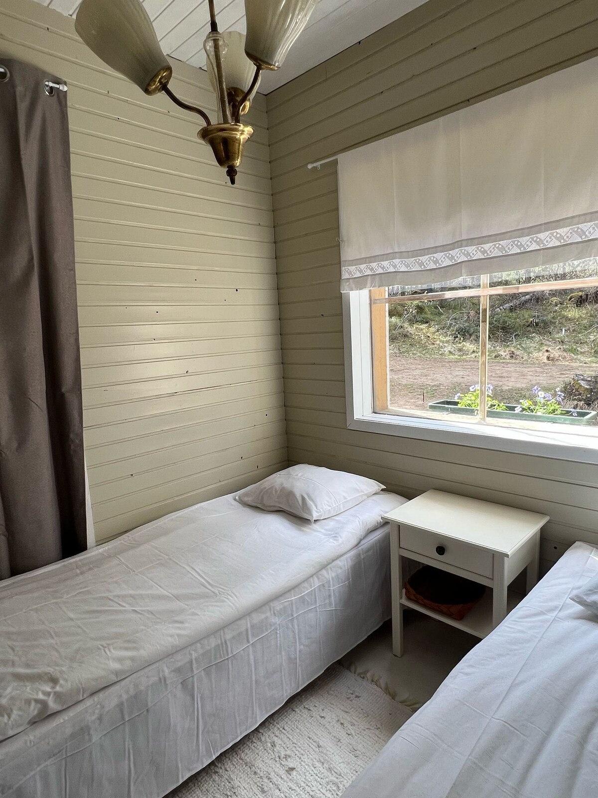 The sleeping area contains two twin beds with white bed linens, positioned beside a small nightstand. Natural light enters through a window with a light-colored blind, offering a view of the outdoors. Soft, neutral tones are complemented by simple decor.