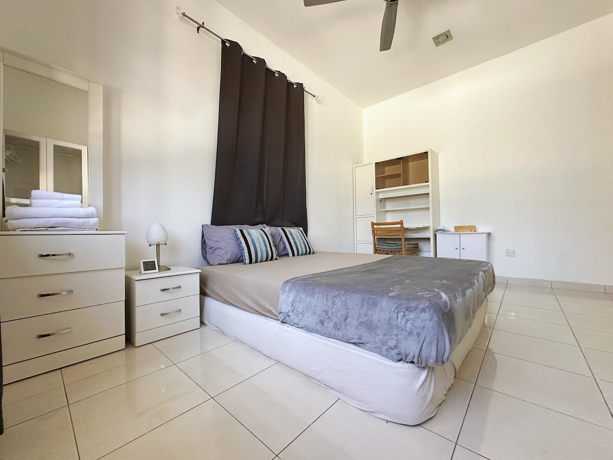 A spacious bedroom is displayed with a king-size bed featuring soft bedding and an accent throw. A light fixture is placed on a bedside table next to the bed. A dresser and a desk with a chair are visible, along with a closet for storage.