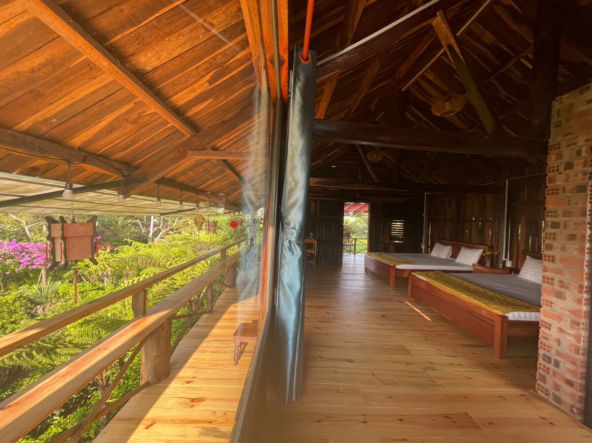 An open space is highlighted by wooden floors and a high ceiling. Large windows offer views of the surrounding greenery. Two sleeping areas are visible, featuring simple bedding and natural light filtering through, creating a serene atmosphere.