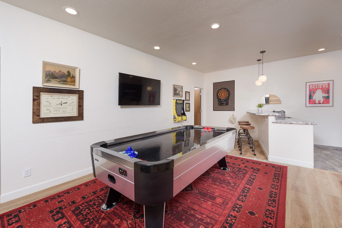 A game room is shown featuring an air hockey table on a decorative red rug. A television is mounted on the wall, and a bar area with stools can be seen in the background. Artwork decorates the walls, contributing to a vibrant atmosphere.