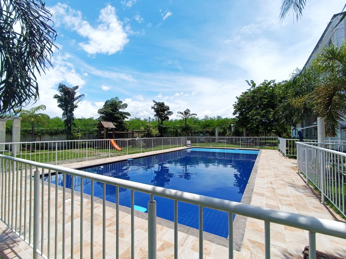A large swimming pool is surrounded by a protective fence, reflecting blue skies and greenery. Relaxing loungers are positioned nearby, and a hammock can be seen in the distance, with lush trees providing shade in the tranquil garden area.