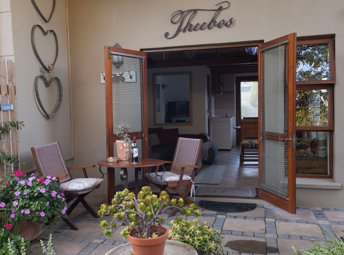 An outdoor patio area is depicted, featuring two folding chairs beside a small round table. A bottle of wine and two glasses sit atop the table. Surrounding the patio, colorful flowers and green plants create a pleasant garden atmosphere. The entrance to the interior space is visible through open double doors.