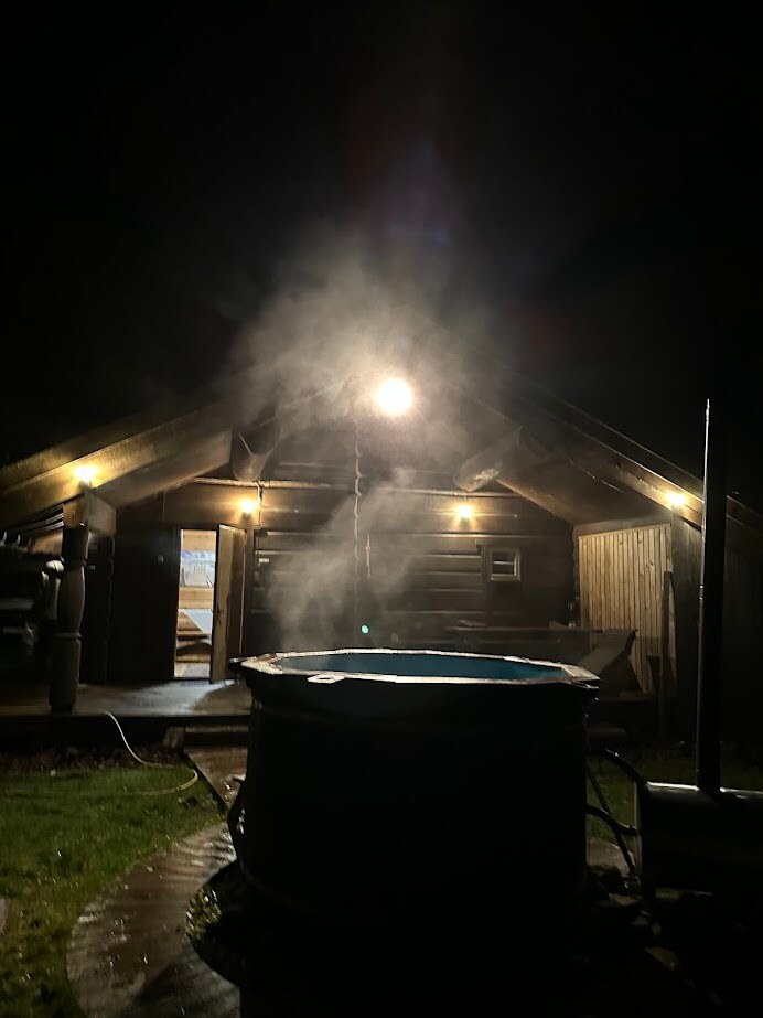 A hot tub emits steam under a dimly lit canopy, creating a serene ambiance. The exterior of a log cabin is visible in the background, showcasing wooden beams and warm indoor lighting. The surrounding area appears lush and inviting, enhancing the tranquil evening atmosphere.