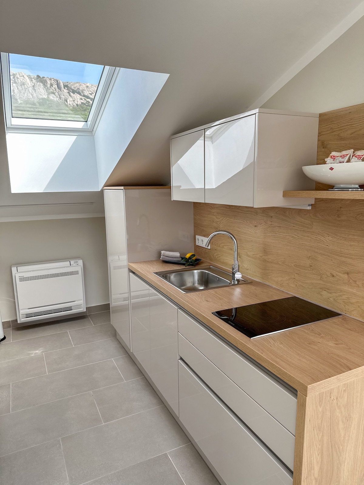 A modern kitchen is displayed with sleek white cabinetry and wooden accents. A stainless steel sink and a ceramic cooktop are present on the countertop. Natural light enters through a skylight, highlighting the minimalist design of the space.