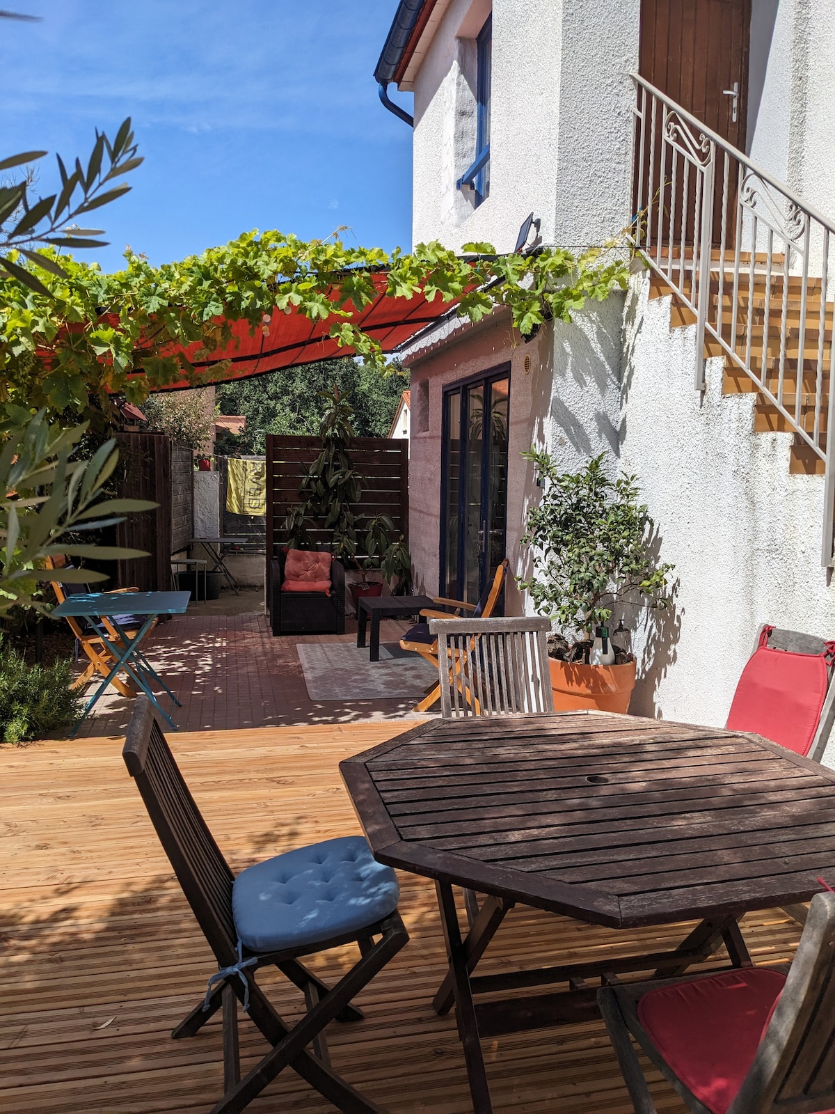 An outdoor patio area features wooden decking surrounded by greenery. A wooden table is complemented by four chairs, while a shaded area provides comfortable seating. Steps lead to the entrance of the adjacent building, and a trellis with vines adds a natural touch.
