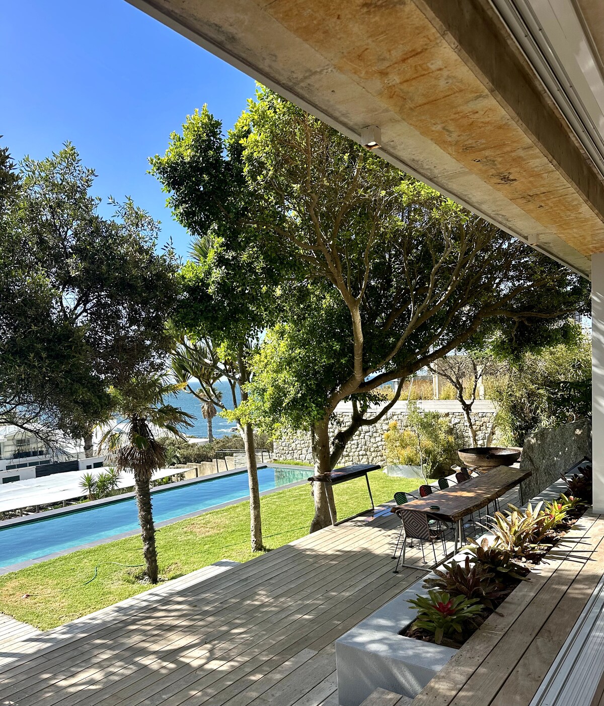 A spacious outdoor area features a sleek swimming pool lined with lush greenery and tall trees. A long dining table with chairs is positioned on a wooden deck, offering a view of the serene landscape and the sea beyond.
