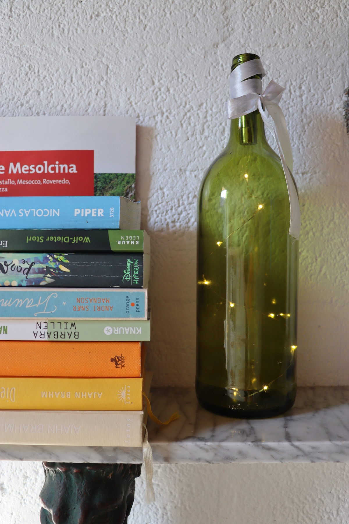 A stack of colorful books is positioned on a marble surface, with a decorative wine bottle beside them. The bottle contains warm string lights, adding a subtle glow. The arrangement creates an inviting atmosphere, ideal for relaxing with a good read in the cozy environment.