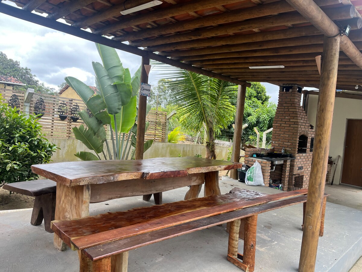 A spacious outdoor dining area features a large wooden table and bench, set under a bamboo-covered structure. Tropical plants are visible in the background, and a barbecue grill is situated nearby, providing a welcoming space for gatherings.