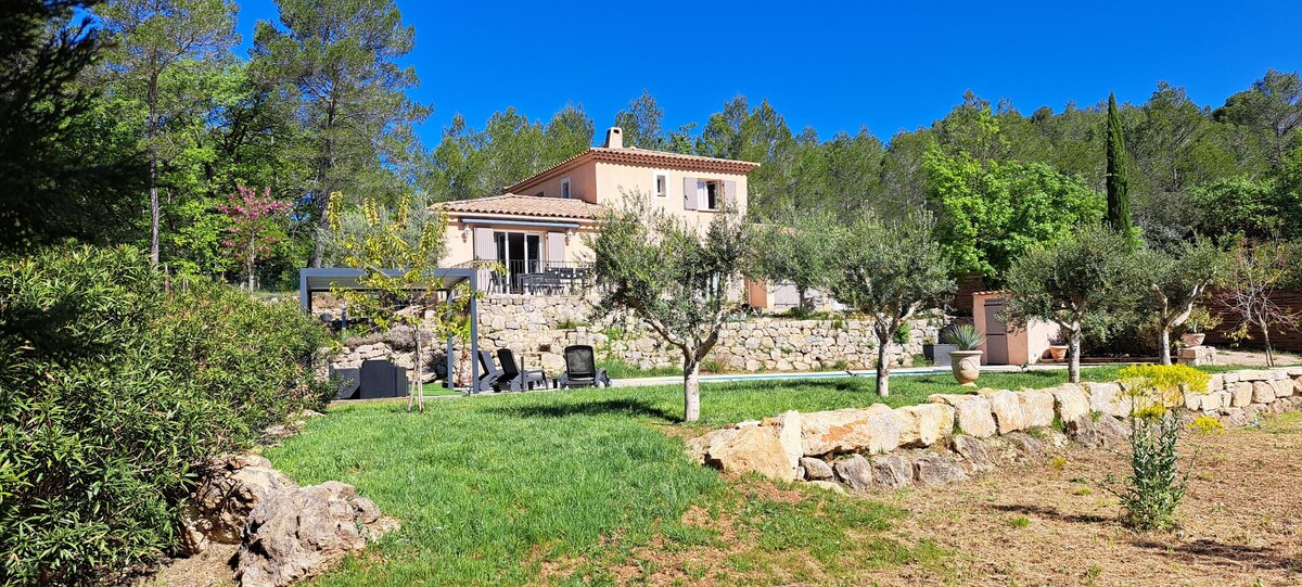 A charming house is set against a backdrop of lush greenery. The outdoor area features well-maintained gardens, stone walls, and a seating arrangement. Sunlight brightens the space, enhancing the natural colors of the trees and plants surrounding the property.