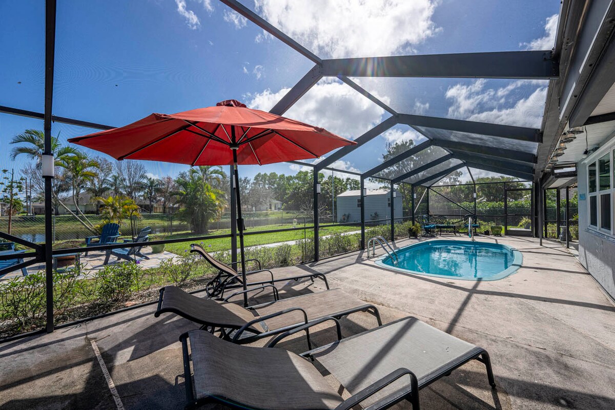 A private pool area is enclosed by a screened lanai, featuring a bright red umbrella above a lounge chair. Several lounge chairs are positioned around the pool, which is surrounded by lush landscaping and offers views of the surrounding greenery.