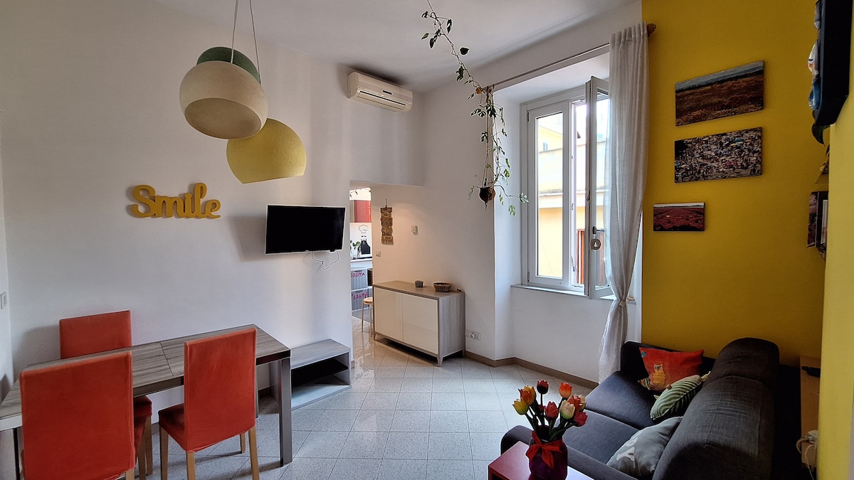 A bright living area features a comfortable gray sofa and a dining table with two red chairs. Large windows allow natural light to fill the space. A television is mounted on the wall opposite a decorative accent wall in sunny yellow, adorned with artwork.