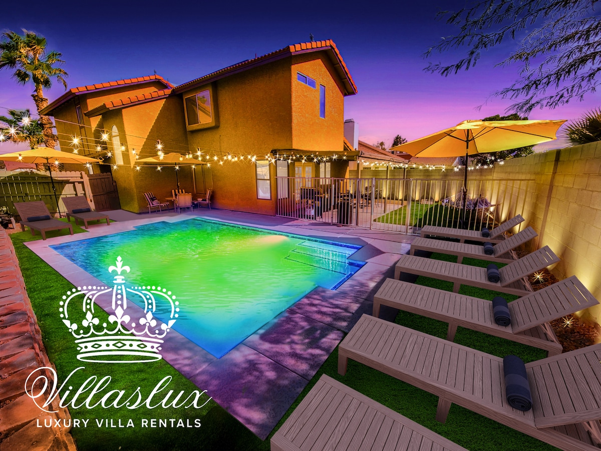 A spacious backyard features a swimming pool with vibrant green and blue hues, surrounded by a decorative fence. Several sun loungers are arranged nearby, with umbrellas providing shade. String lights illuminate the area, creating a warm ambiance as evening approaches.