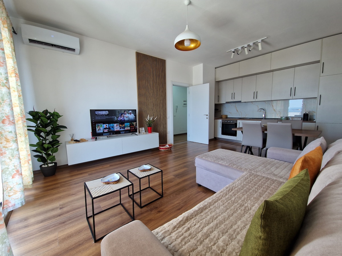 A modern living space is presented, featuring a comfortable sectional sofa and a pair of small coffee tables. A large television is mounted on the wall, with soft lighting provided by a ceiling fixture. The room includes a kitchenette area, and a potted plant adds a touch of greenery.