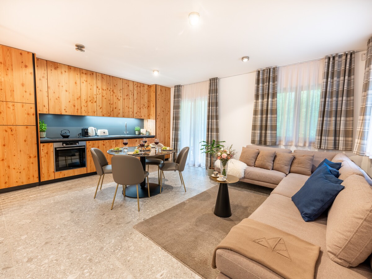An open-plan living area includes a modern kitchen featuring wooden cabinets and appliances. A round dining table is surrounded by four chairs. A cozy sofa with decorative cushions faces the table, while soft natural light enters through large windows adorned with checkered curtains.