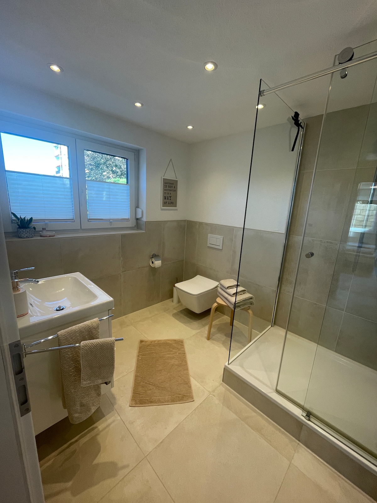 A modern bathroom is equipped with a walk-in shower featuring a glass enclosure. Soft lighting illuminates the neutral-colored tiles. A compact sink is positioned next to a small chair, while a bath mat is placed on the floor. Natural light filters through a nearby window.