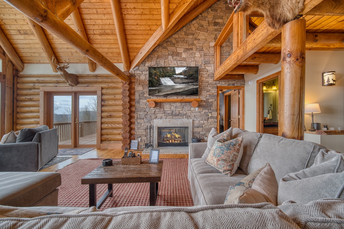 A spacious living room showcases a stone fireplace framed by logs and a large mountain-view window. Comfortable seating is arranged around a rustic coffee table, with a flat-screen TV mounted above the fireplace. Natural wood accents and warm tones create a welcoming ambiance.