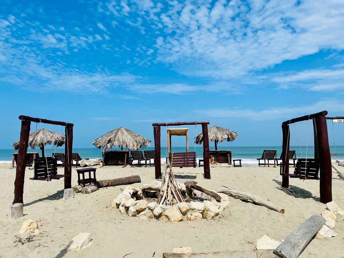 A sandy beach area is highlighted with several seating options, including wooden chairs and swing sets. Thatched-roof huts provide shaded spots. A circular arrangement of stones indicates a bonfire area, and the calm ocean waves are visible in the background under a clear blue sky.