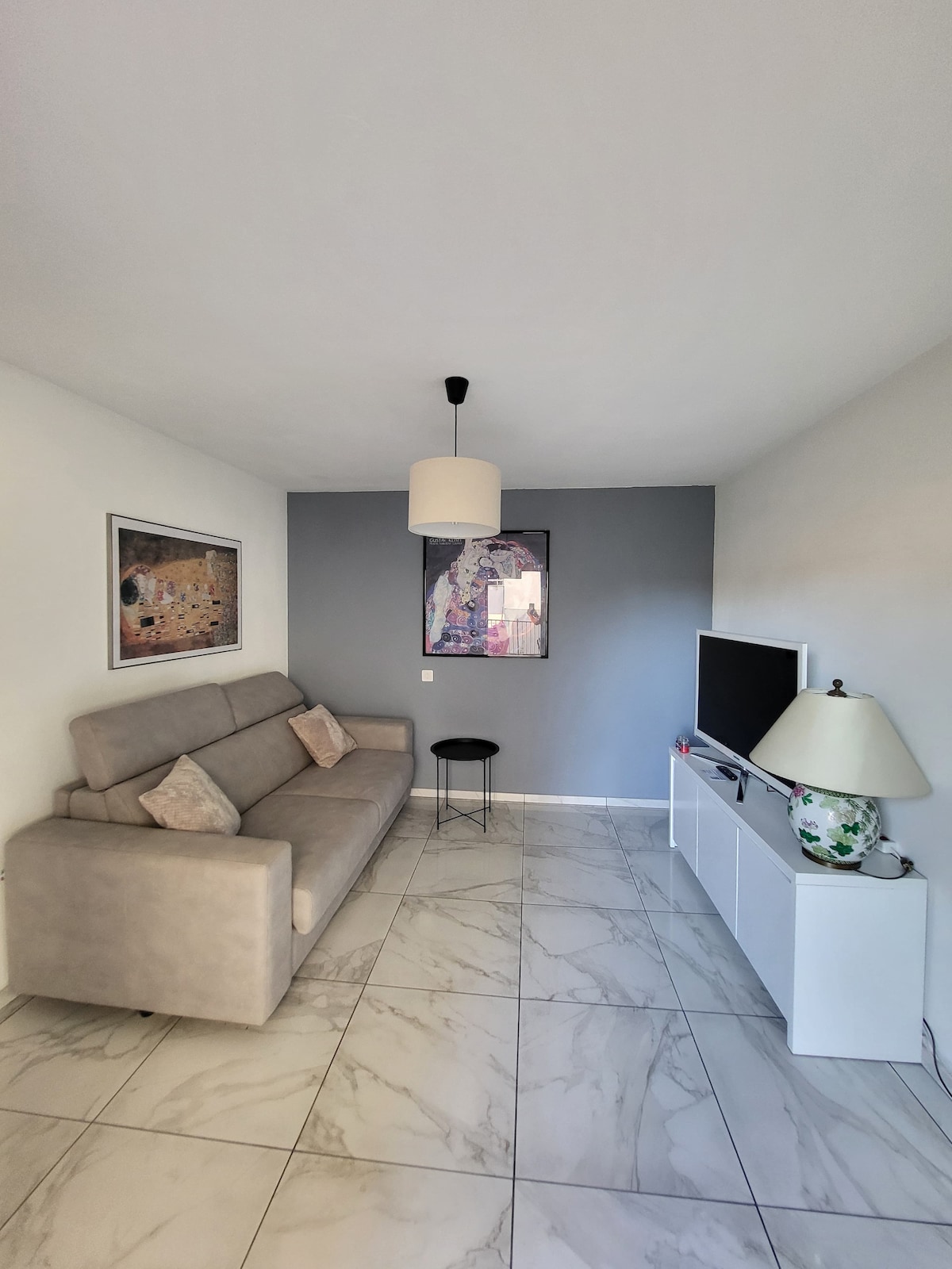 A light and airy living space features a grey accent wall and a comfortable sofa complemented by two decorative pillows. A minimalist side table stands beside the sofa, while a flat-screen TV is mounted on a white media console. Artwork is displayed on the wall opposite the sofa.