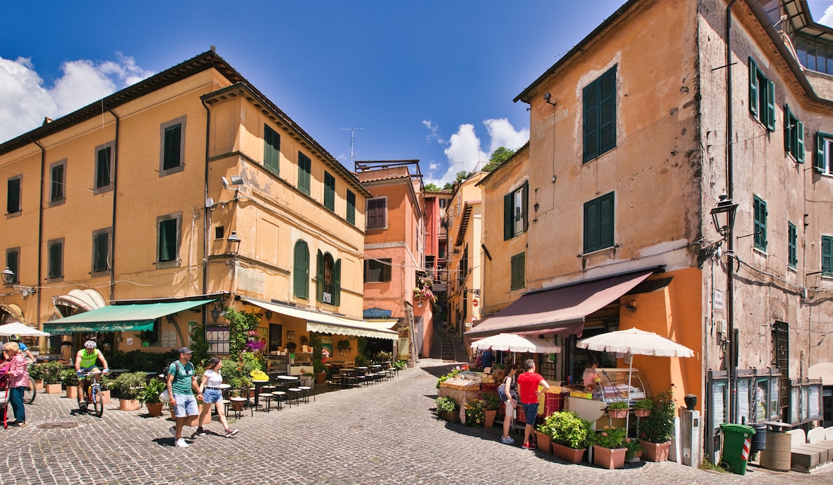 A vibrant village square is displayed with charming buildings featuring pastel-colored facades and green shutters. Flower-filled planters line the cobblestone streets. Outdoor seating areas are seen, accompanied by market stalls showcasing fresh produce. Visitors are engaged in leisurely activities under a clear blue sky.