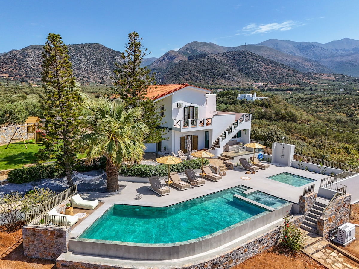 A spacious villa with a bright exterior is surrounded by lush greenery and mountains. An inviting swimming pool is centrally located, flanked by sun loungers. Nearby, a shaded area offers seating under a pergola, while a playground is visible in the background.