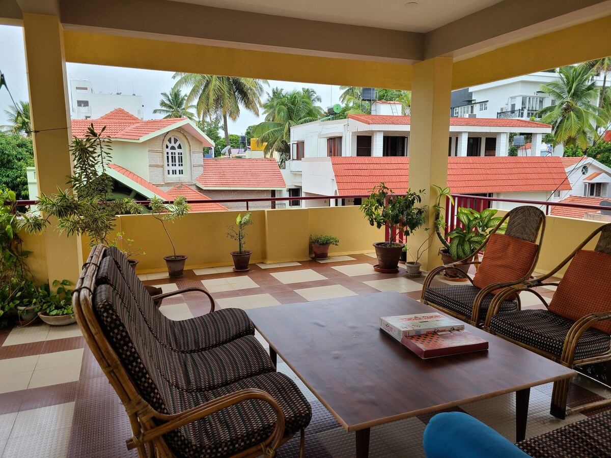 An outdoor terrace is shown with comfortable seating arrangements that include multiple chairs and a spacious table. Potted plants add greenery to the space, while neighboring buildings with red roofs and palm trees are visible in the background.