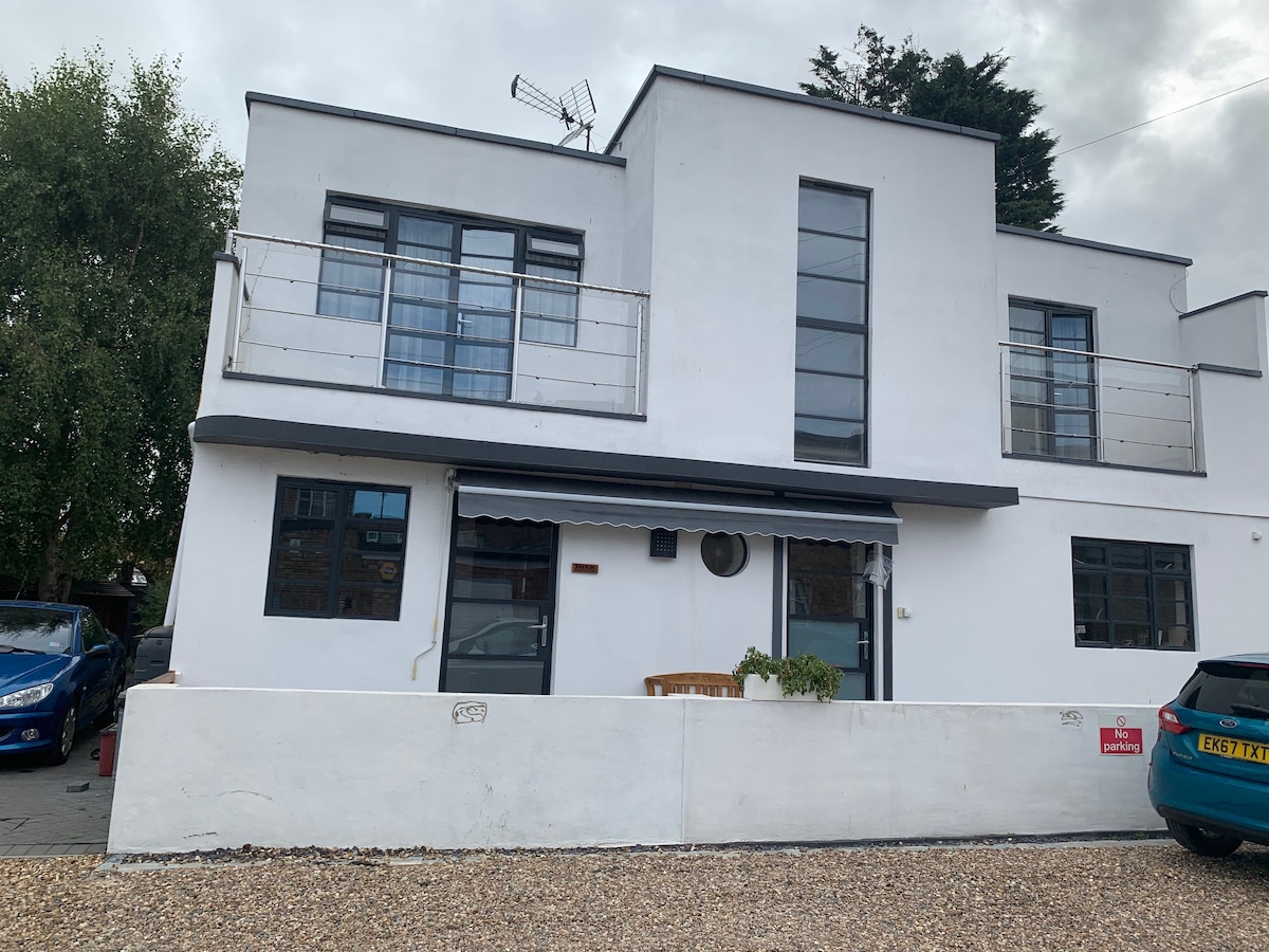 The exterior of the modern Annexe is presented, showcasing a white façade with large windows that allow natural light. A small balcony is visible above, accompanied by a well-maintained gravel driveway featuring designated parking spaces for guests.