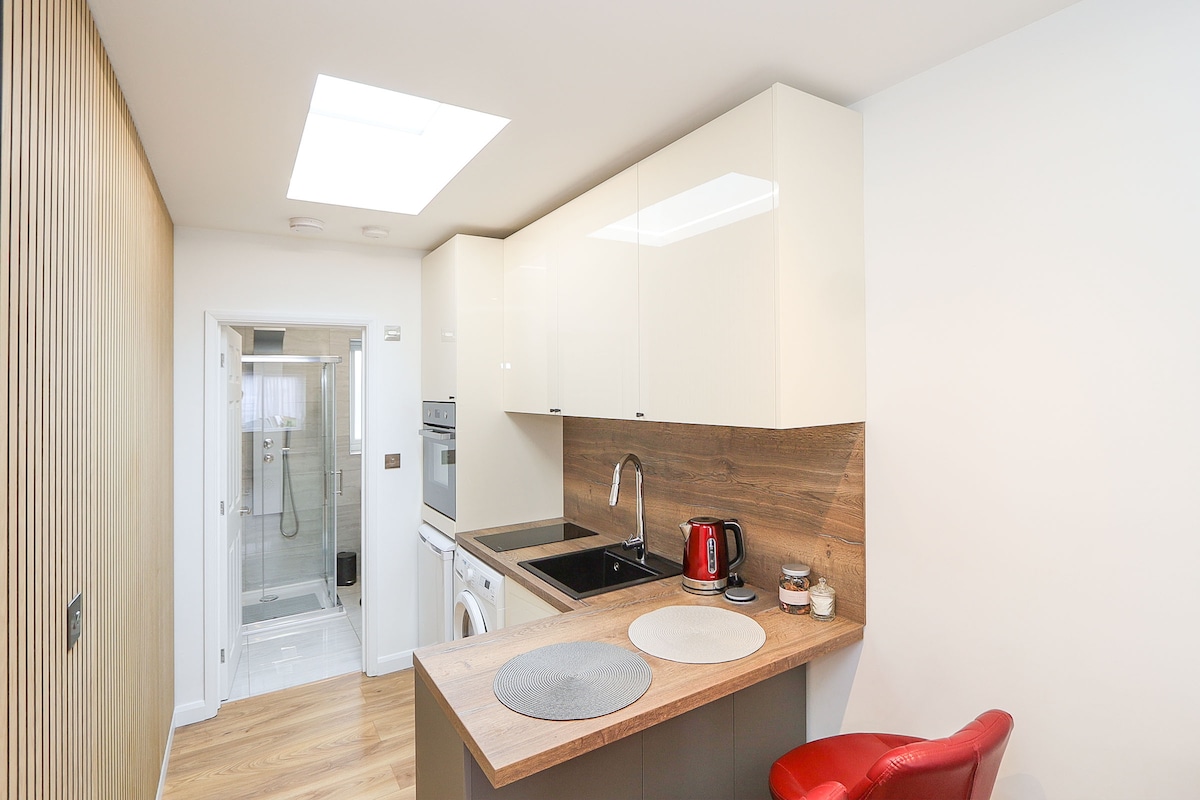 A compact kitchen area features modern cabinetry with an integrated oven and a seamless stovetop. A kettle and additional kitchen essentials are positioned on the counter. The space is illuminated by natural light streaming through a skylight above, enhancing the warm wooden tones of the flooring.