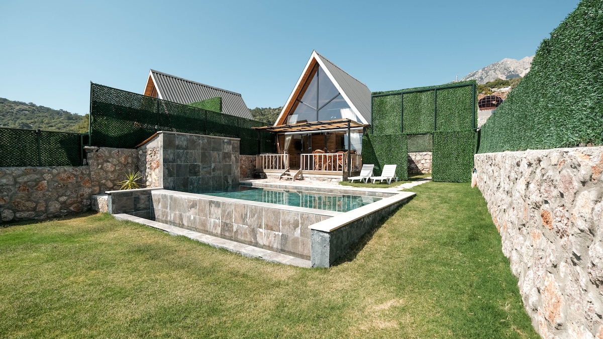 A modern villa is surrounded by lush greenery and stone walls, featuring a private pool in front. Comfortable loungers are placed beside the pool, inviting relaxation. The building's design incorporates large windows and a spacious deck, creating an openness to the outdoors.