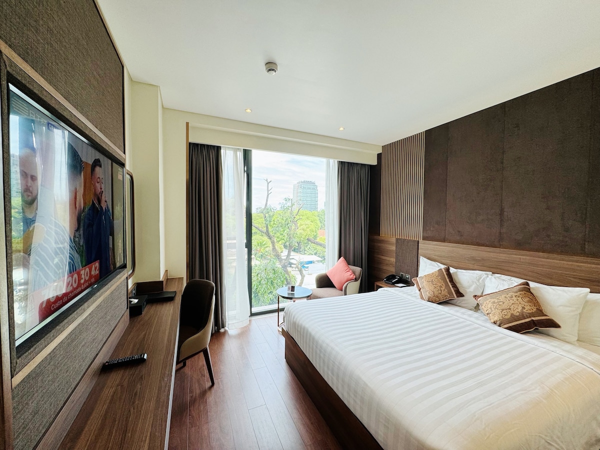 A contemporary hotel room features a spacious bed with decorative pillows and a flat-screen TV mounted on the wall. A work desk and chair are positioned near large windows, offering natural light and views of the surrounding area. Dark curtains frame the windows.
