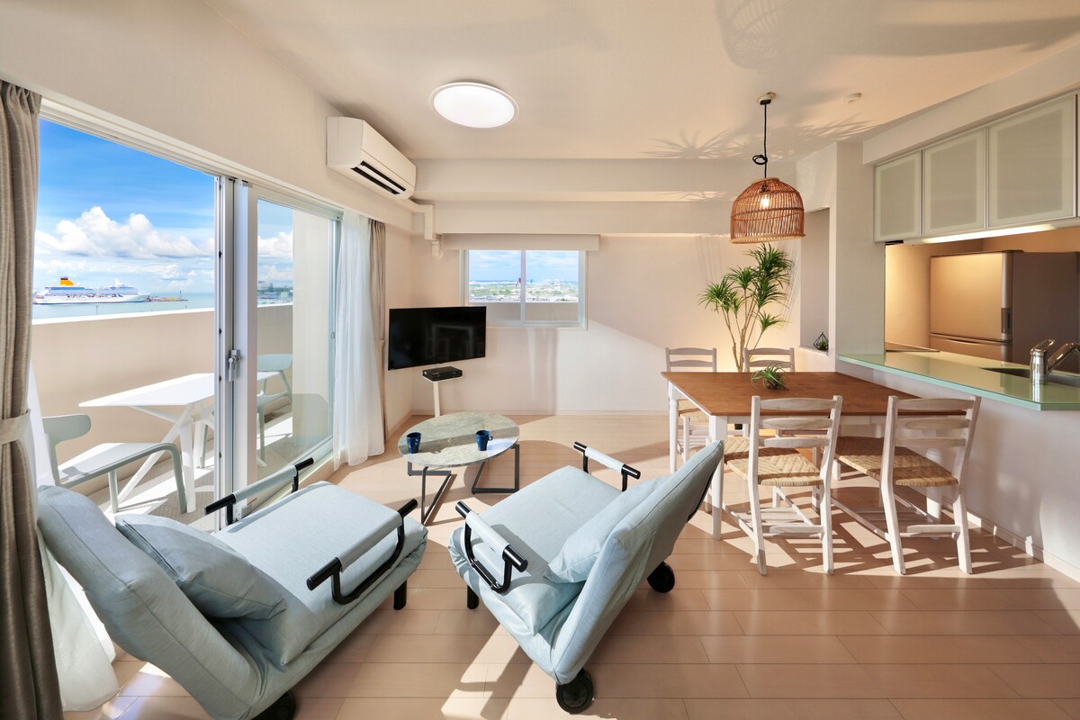 A bright living area is highlighted by large windows that offer views of the outdoors. Two blue lounge chairs are positioned nearby, with a dining table and chairs visible in the background, alongside an open kitchen space. Sunlight fills the room, creating a fresh ambiance.