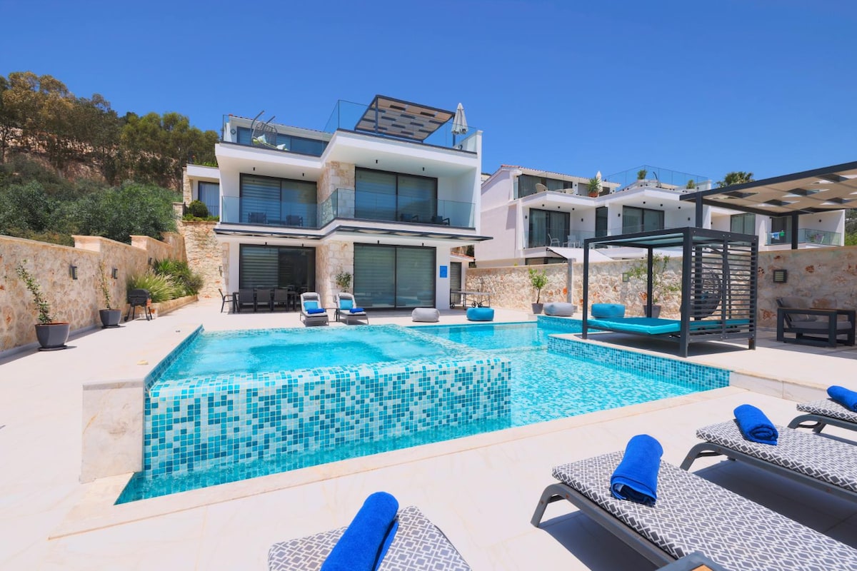 A modern villa with a striking facade showcases a spacious pool area with a built-in jacuzzi. Sun loungers are placed by the clear water, and a shaded seating area offers a stylish retreat. Glass doors provide access to the indoor spaces, enhancing the overall contemporary design.