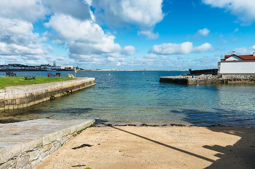 The image captures a tranquil coastal scene, featuring calm blue waters meeting a sandy shoreline. Lush greenery and rocky formations are visible in the foreground, with distant views of the town and cloudy sky enhancing the serene atmosphere.