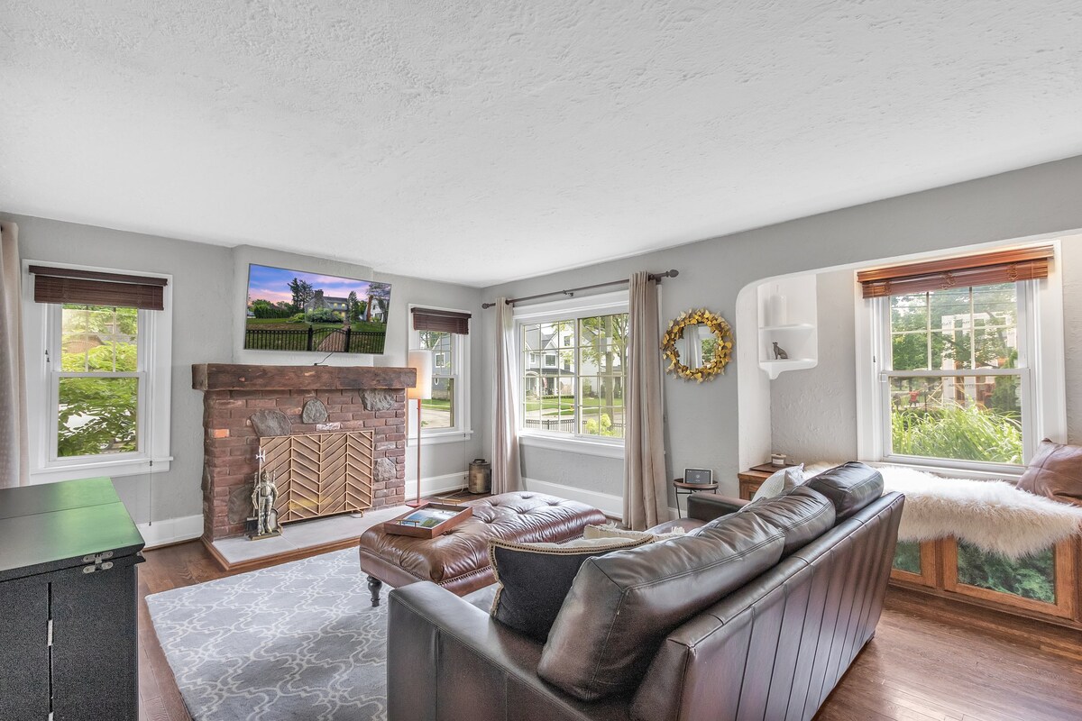 A bright and airy living room is displayed, featuring a cozy leather sofa and a large area rug. A stone fireplace serves as a focal point, while large windows allow natural light to fill the space. A television is mounted above the fireplace, enhancing the comfort of the area.