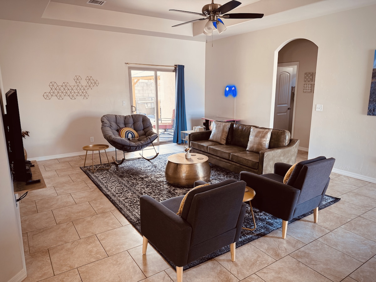 A spacious living area features a large sectional sofa and two accent chairs, all set against a light-colored wall. A circular coffee table sits in the center, while a sliding glass door leads to an outdoor space. Decorative elements, such as a wall-mounted light and artwork, add character.