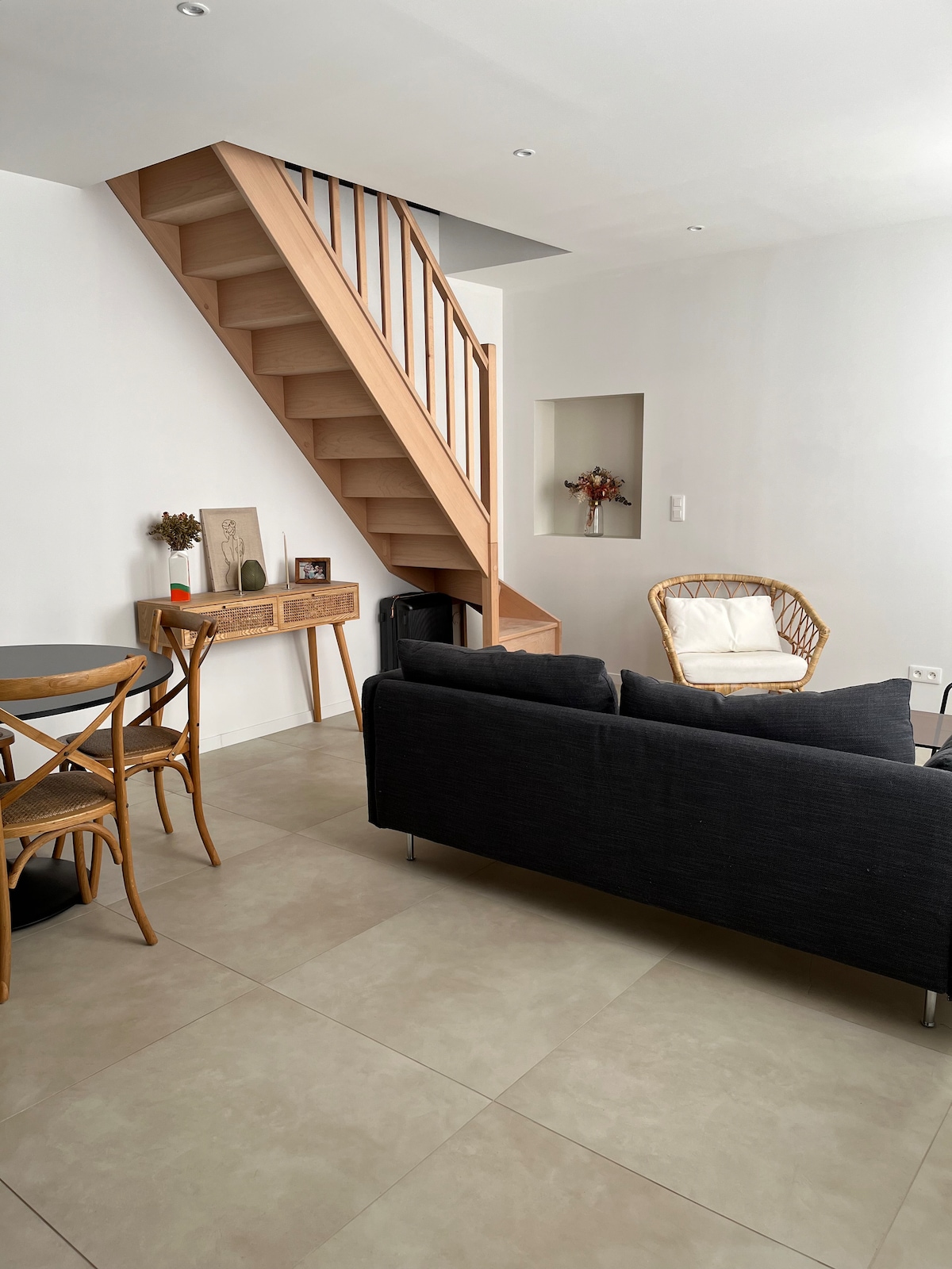 A modern living space features a dark sofa facing a small wooden table and chairs. A staircase with wooden steps leads to an upper level. A decorative piece and a small vase with dried flowers are displayed on a console table against the wall.