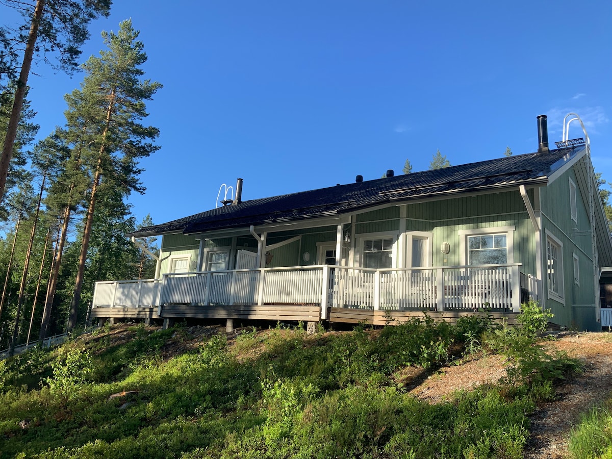 A green wooden structure is surrounded by trees and features a spacious deck with white railings. The angled roof is covered with dark tiles, and large windows provide views of the surrounding nature. The building is situated on a gentle slope, enhancing its scenic setting.