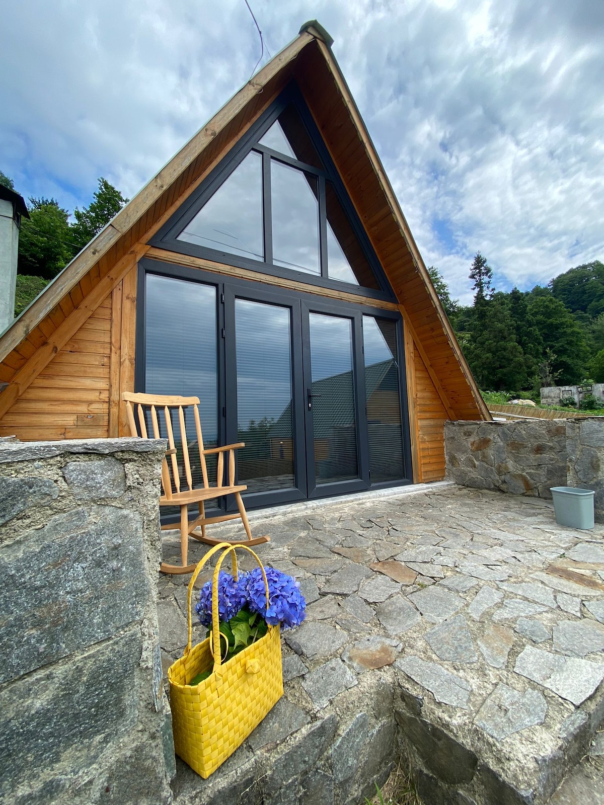 The exterior of the house is showcased with a wooden triangular design, featuring large glass doors that open to the outside. A rocking chair is placed on the stone patio, accompanied by a yellow basket filled with blue flowers, creating a welcoming outdoor space.