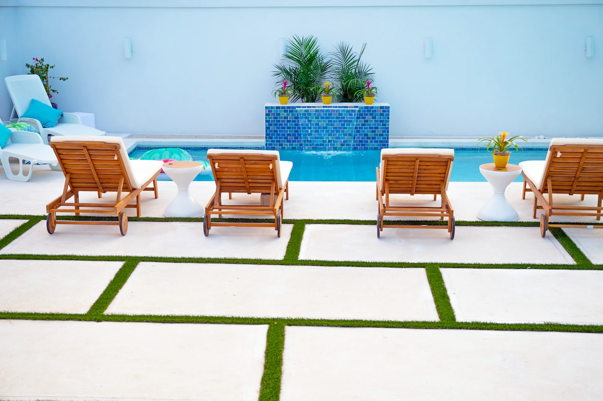 A private pool area features several brown wooden lounge chairs arranged neatly on light-colored tiles, separated by sections of greenery. A small water feature with blue mosaic tiles is visible in the background, complemented by colorful potted plants.