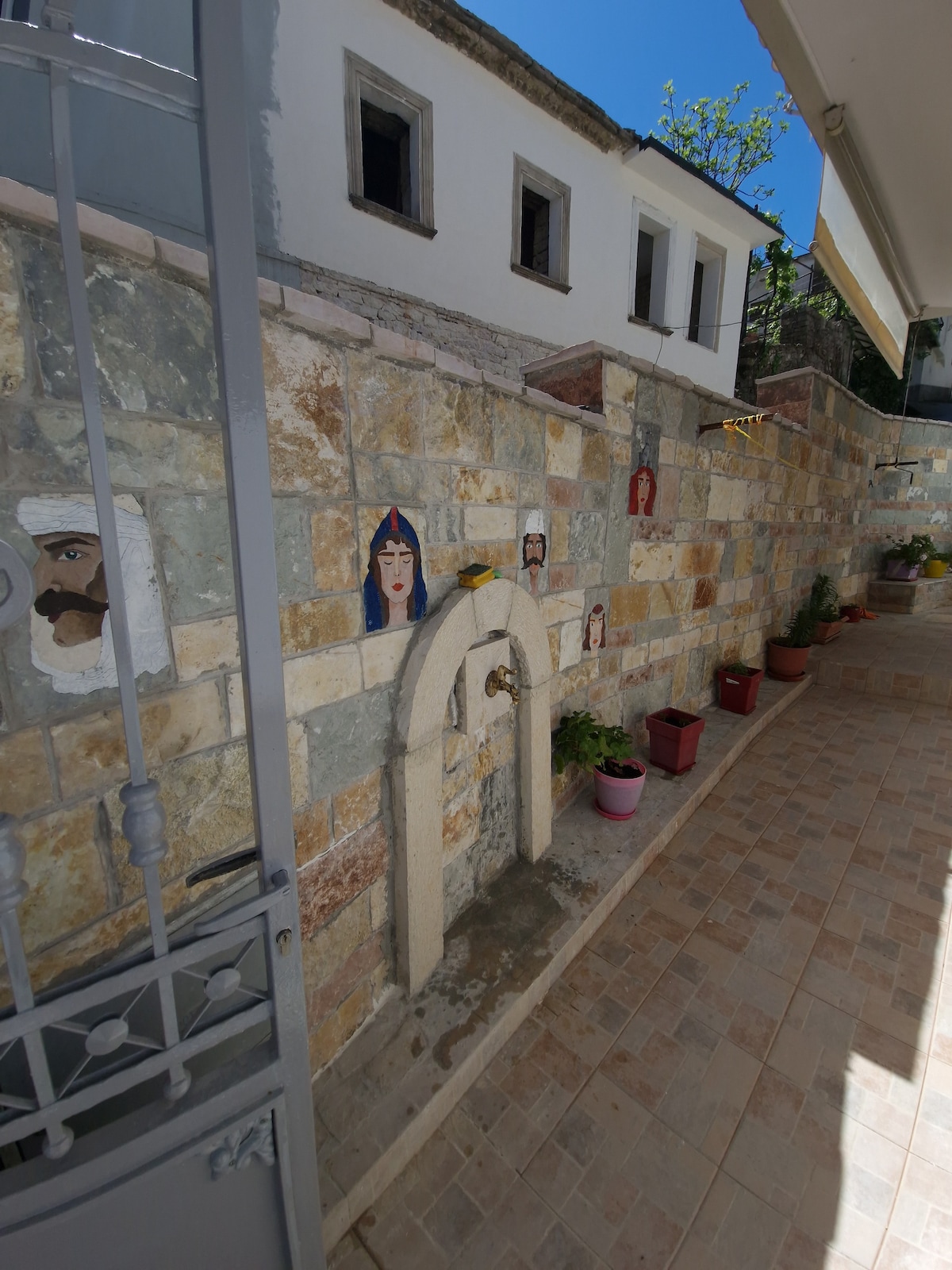 A stone wall adorned with artwork and potted plants creates a charming outdoor path. Sunlight is seen casting shadows on the pavement, and a small archway is featured prominently among the decorations. The overall setting suggests a peaceful, welcoming entrance.